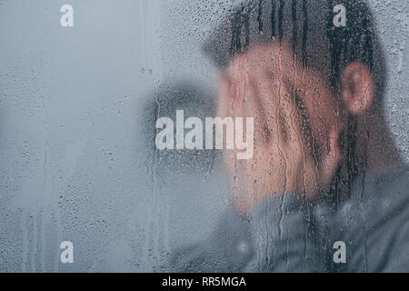 selective focus of raindrops on window with man covering face and crying on background Stock Photo