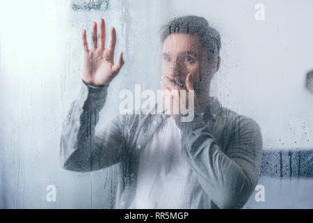 upset man covering mouth with hand and touching window with raindrops Stock Photo