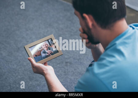 back view of adult man grieving while looking at photo frame with old man at home Stock Photo