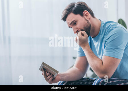 depressed man sitting, covering mouth with hand and looking at photo frame at home Stock Photo