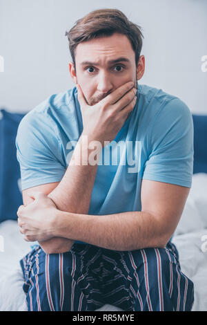 stressed man sitting on bed, looking at camera and covering mouth with hand at home Stock Photo