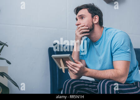 upset man sitting on bed, covering mouth and holding photo frame at home Stock Photo