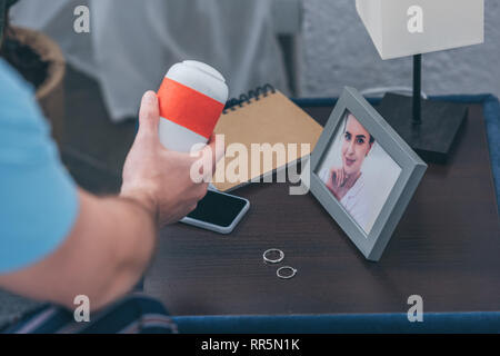 cropped view of man holding funeral urn near picture of woman and wedding rings on table Stock Photo