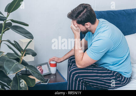 man photo holding funeral urn, covering face with hand and grieving near photo frame with picture of woman and wedding rings Stock Photo