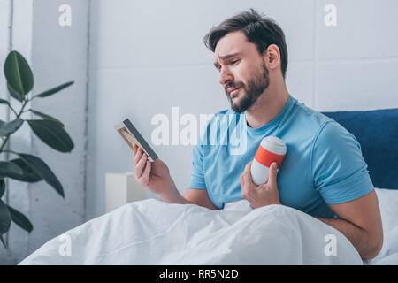 man lying in bed, crying and holding photo frame with funeral urn Stock Photo