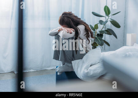 grieving woman sitting, covering face with hands and crying in bedroom Stock Photo