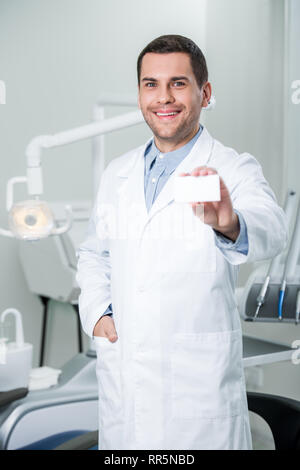 cheerful dentist standing with hand in pocket and holding blank card Stock Photo