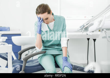exhausted dentist holding head while sitting on chair in dental clinic Stock Photo
