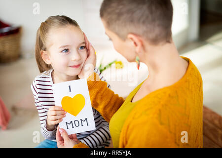 Young female cancer patient thanking her adorable young daughter for homemade I LOVE MOM greeting card. Family celebration concept. Happy Mother's Day Stock Photo