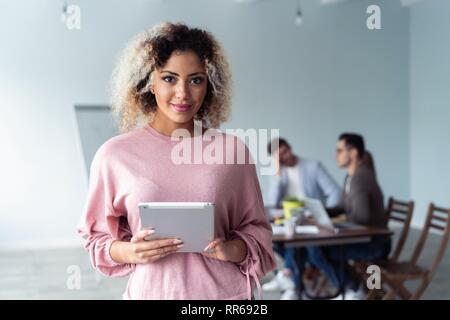 Business woman standing in foreground with a tablet in her hands, her co-workers discussing business matters in the background. Stock Photo