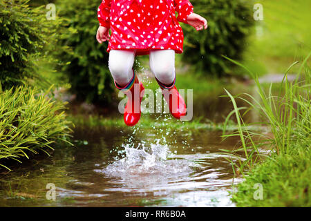 Kid playing in the rain in autumn park. Child jumping in muddy puddle on rainy fall day. Little girl in rain boots and red jacket outdoors in heavy sh Stock Photo