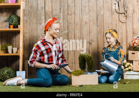 mother holding shovel near daughter watering plant while sitting on grass near wooden fence Stock Photo