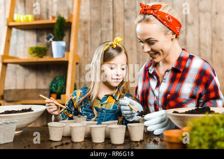 cheerful mother holding small shovel and putting ground in empty paper cups near cute daughter Stock Photo