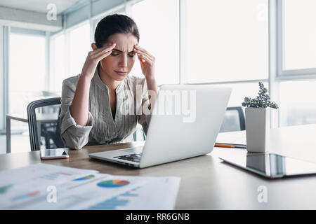 Young dark-haired businesswoman wearing striped shirt is sitting in her office, working on laptop, massaging temples to forget about constant headache Stock Photo
