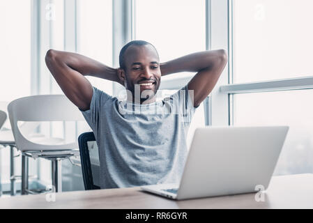 Feeling joy. Cheerful young african american businessman is reclining on chair and holding his hands behind head while looking on the screen with wide Stock Photo