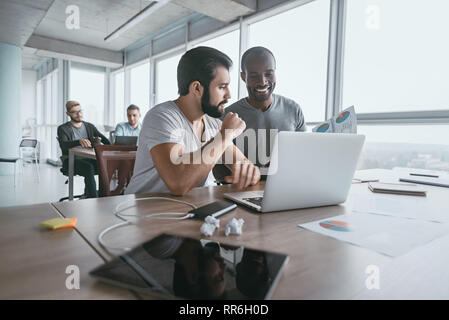 Two young businessmen using laptop and discussing new project at office, developing strategy for online business, explaining sharing ideas, preparing presentation, having brainstorming session Stock Photo