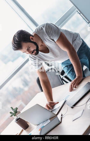 Young man in casual wear standing over his office table, using his laptop. Working papers and gadgets lie on desk. Concept of decision making Stock Photo
