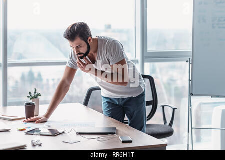 Young man in casual wear standing over his office table, thinking about business and planning. Working papers and gadgets lie on desk. Concept of decision making Stock Photo