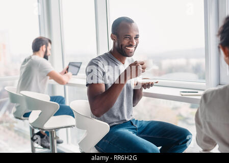 Portrait of multicultural employees dressed in casual wear, laughing while enjoying coffee break during working process in modern office interior, behind glass wall Stock Photo