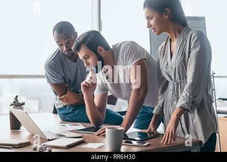Group of attractive multiracial businesspeople working together in creative office while standing near the wooden desk with a laptop on it, friendly multi-ethnic business team looking on the screen, discussing business issues at meeting Stock Photo