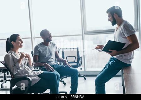 Group of attractive multiracial laughing businesspeople talking and joking sitting on chairs in light modern office, friendly multi-ethnic business team having fun, discussing business issues at meeting Stock Photo