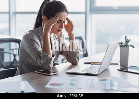 Young dark-haired businesswoman wearing striped shirt is sitting in her office, working on laptop, massaging temples to forget about constant headaches, about noisy office giving a migraine. She is trying to relieve stress and chronic pain Stock Photo