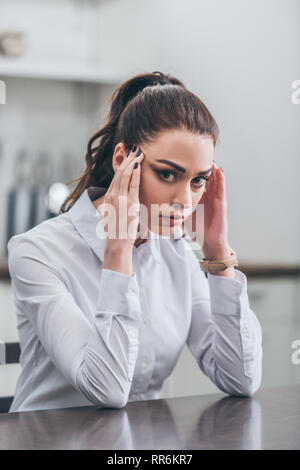 upset woman in white blouse sitting at table in kitchen and holding hands on temples, grieving disorder concept Stock Photo