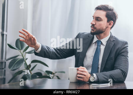 sad man in gray suit sitting at wooden table with smartphone holding white cap and looking away at home, grieving disorder concept Stock Photo