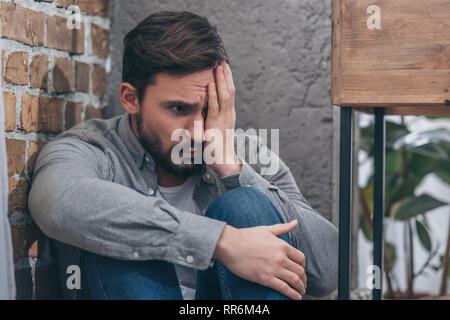 upset man sitting in corner, covering eye with hand on brown textured background in room, grieving disorder concept Stock Photo