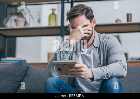 upset man in gray shirt covering face with hand and holding photo frame at home, grieving disorder concept Stock Photo