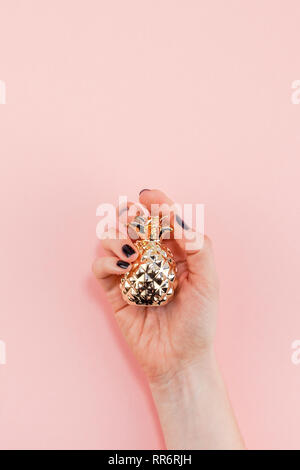 Creative top view flat lay of woman hand with black manicure holding small golden pineapple with copy space on pink paper background in minimalism sty Stock Photo