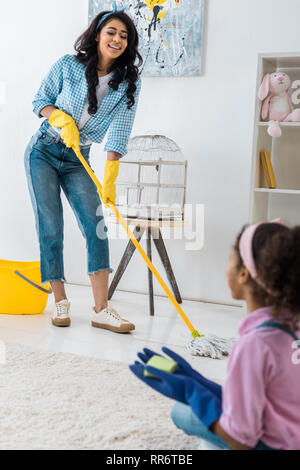 smiling african american woman washing floor with mop while daughter sitting on carpet Stock Photo