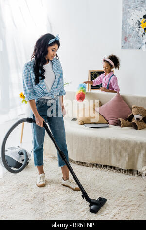 attractive african american woman cleaning carpet with vacuum cleaner while cute daughter dusting sofa Stock Photo