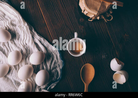 Yolk in cup next to whole eggs and shells on wooden table, top view. Ingredients for baking in rustic style. Stock Photo