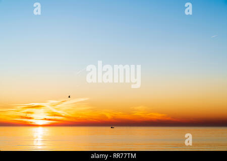 Sunrise, sunup, over the sea, English Channel, at Viking Bay Broadstairs. Thin band of cloud and orange sky on horizon with clear blue sky above. Single boat silhouette on sea. Seagull flying over the sea. Stock Photo