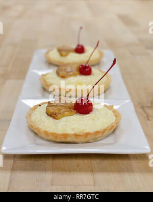 Three custard tarts in a row on white square plates.  Garnished with glazed pineapple and cherries.  Wooden background. Stock Photo