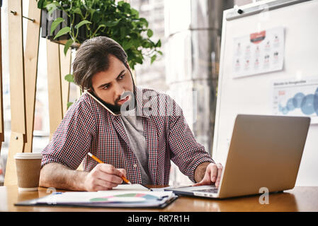 Young serious businessman in casual wear is talking on the phone, while sitting in office in front of laptop, using it, making notes in his notebook. Freelancer analyzes data, develops development strategy. Business education for adults. Stock Photo