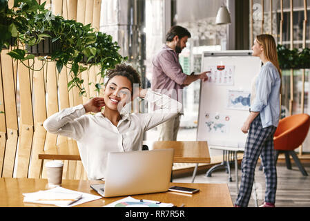 Young business woman relaxing her neck and shoulders to relieve stress at the office, tilting her head to one side smiling and looking away. Co-workers standing near whiteboard in the background. Stock Photo