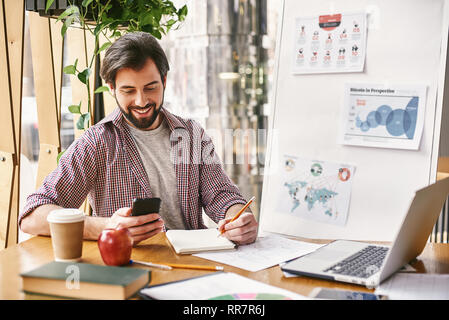 Young cheerful businessman in casual wear is looking on the phone, while sitting in office in front of laptop, using it, making notes on chart. Freelancer analyzes data, develops development strategy. Business education for adults. Notebooks, planers, an apple and a cup of coffee are on the desk. Stock Photo