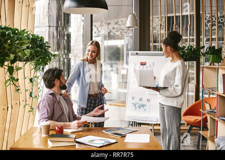 Attractive young woman standing near whiteboard with a laptop in her hands and looking to it. Another co-workers are talking to each other, looking with a smile. Concept of success Stock Photo