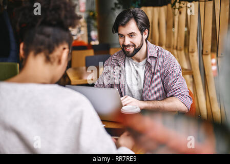 Two cheerful colleagues in office wearing casual clothes co-working while using laptop computer. Bearded dark-haired man looking on his female colleague with a smile, while she is focused on her work. Stock Photo