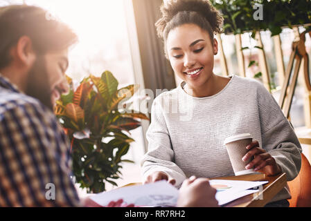 Two cheerful colleagues discussing new ideas sitting over coffee table. Bearded caucasian man and attractive african american woman look on charts. Coffee, working papers and pencils are on the desk. Casual style. Concept of success Stock Photo