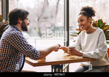 Side view portrait of two successful business people, man and woman, smiling and shaking hands across table during meeting in cafe. African american woman and caucasian dark-haired man Stock Photo