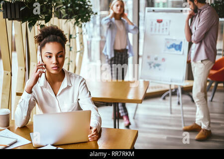 Portrait of serious young woman sitting at her desk, talking on mobile phone. Concentrated business woman talking with client over smart phone in office and looking aside. Woman working at desk in modern office. Her notebook and coffee are on the table. Colleagues in the background. Stock Photo