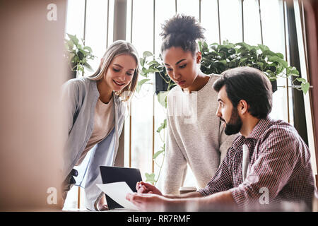 Three cheerful colleagues discussing new ideas in office. Bearded man explains his suggestions, while women listen to him attentively. A laptop computer, charts and coffee are on the desk. Casual style. Concept of success Stock Photo