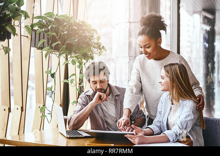 Three cheerful colleagues discussing new ideas in office. Bearded man looks on the chart, while women talk to him. A laptop computer, notebook and pencils are on the desk. Casual style. Concept of success Stock Photo