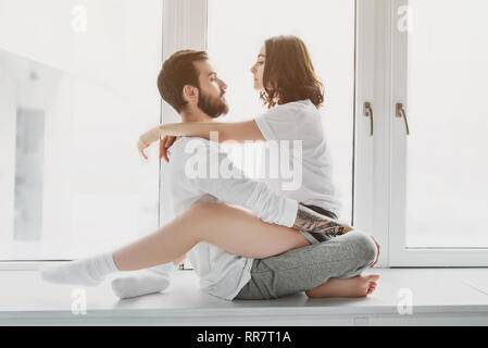 beautiful young couple looking at each other, hugging and sitting on window sill at home Stock Photo