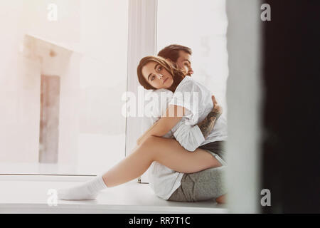 beautiful young romantic couple hugging and sitting on window sill at home with copy space Stock Photo