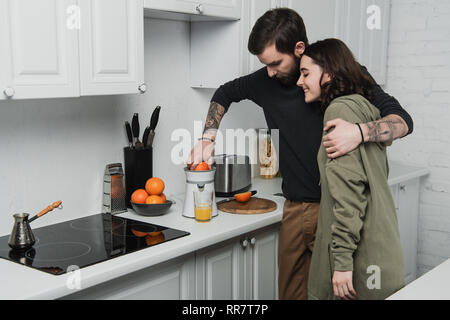 beautiful young couple making orange juice and hugging during breakfast in kitchen Stock Photo