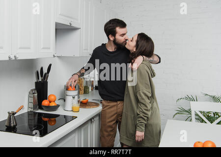 beautiful young couple making orange juice, hugging and kissing during breakfast in kitchen Stock Photo